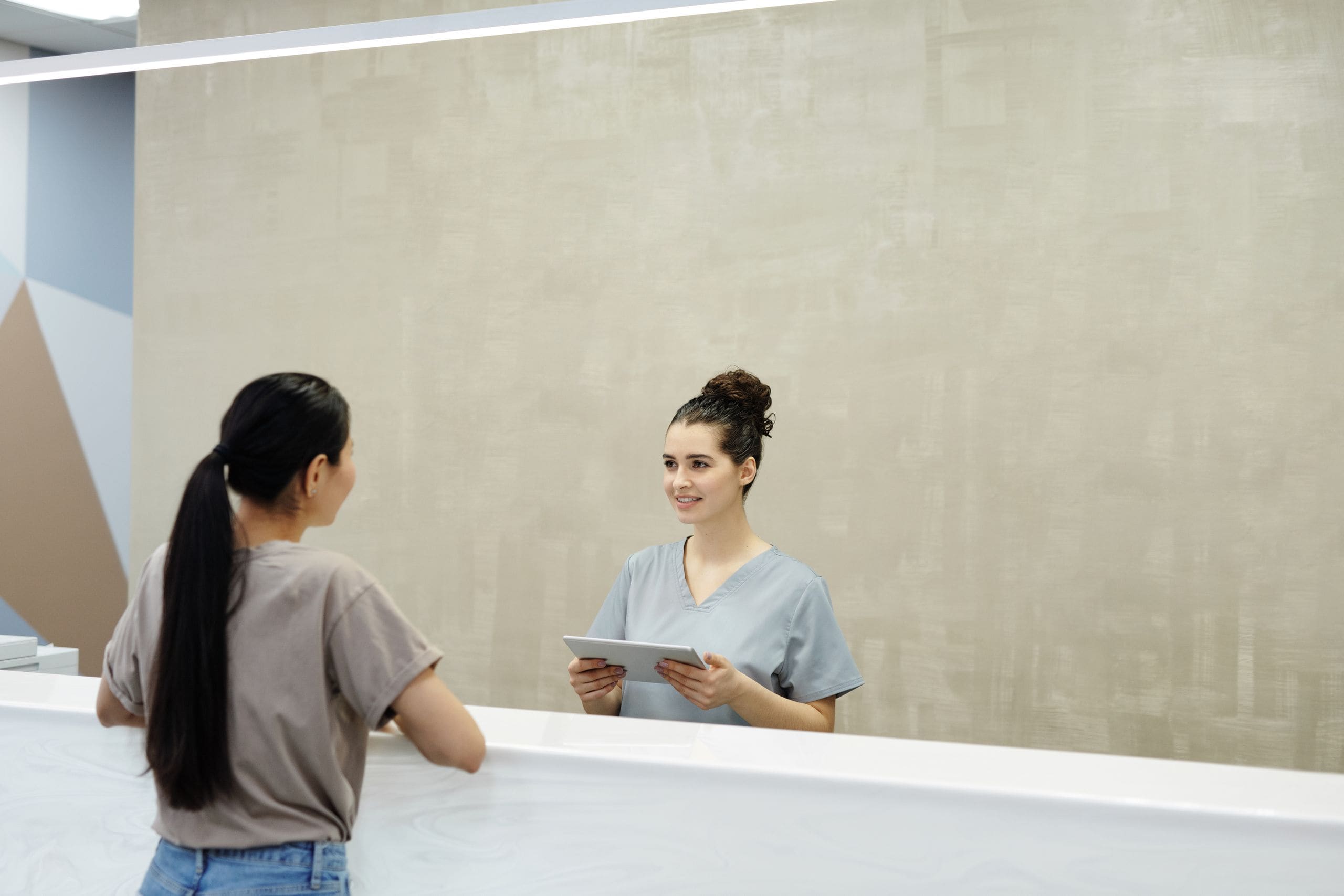 A clinic receptionist with a tablet assisting a patient at the front desk