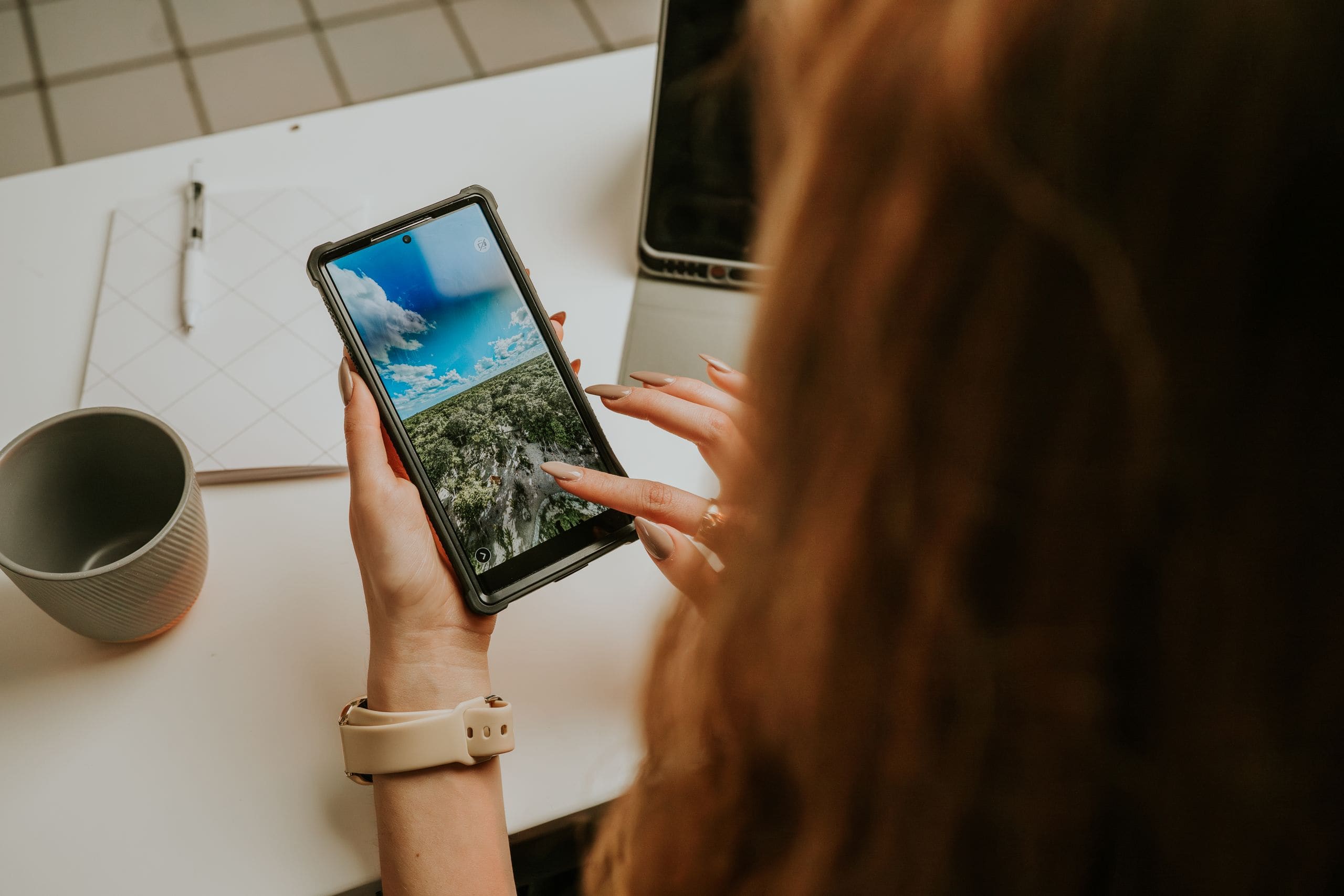 A marketer at her desk choosing a photo on her phone, notebook and coffee beside her