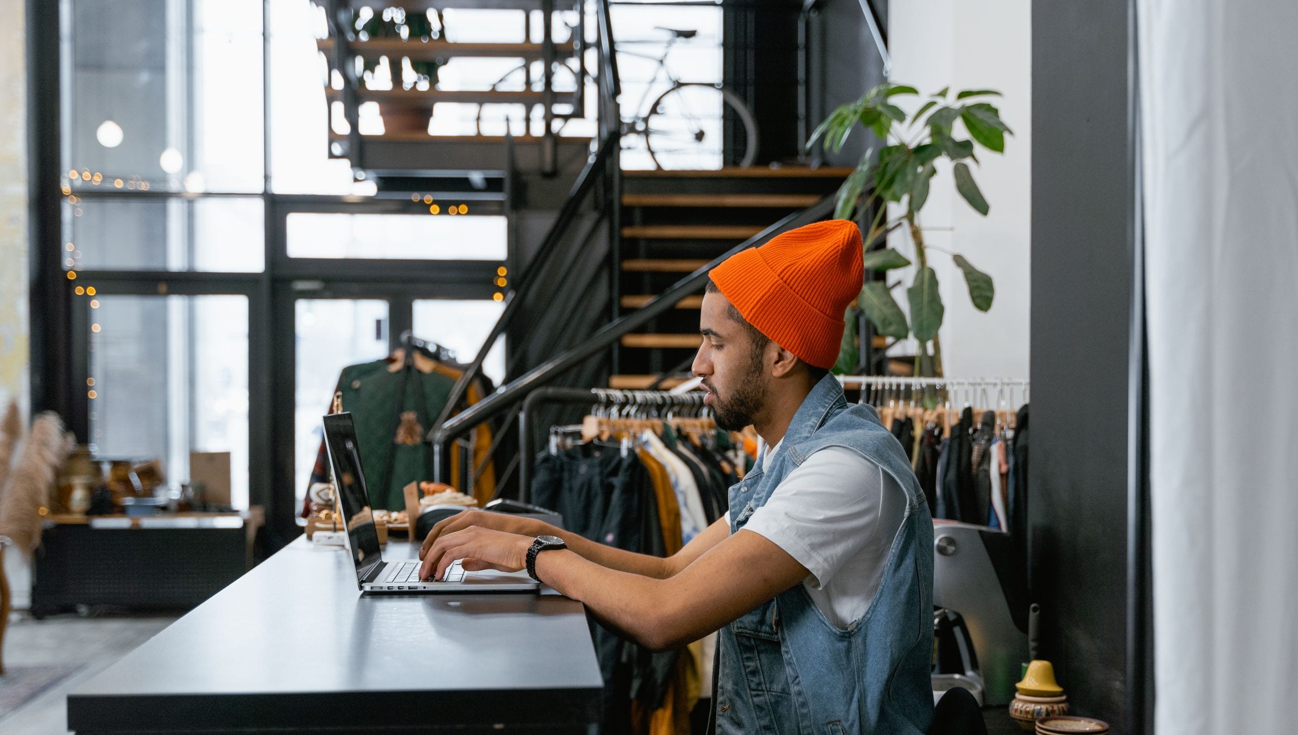 A shop owner in an orange beanie typing on a laptop at the counter of his clothing store